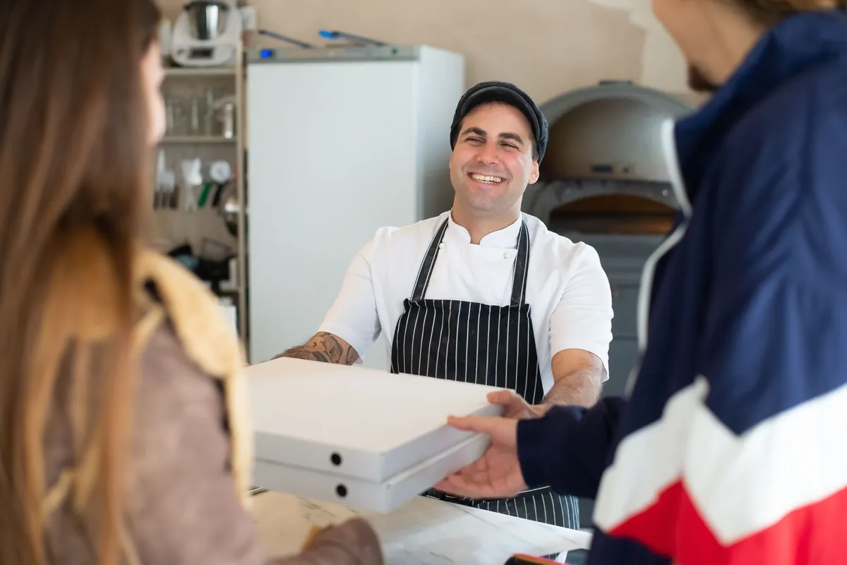 Smiling pizzeria owner in a cap and striped apron handing takeout pizza boxes to customers across the counter